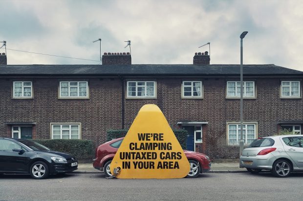 Image of car parked in a street with a giant clamp on it which says "We're clamping untaxed cars in your area"