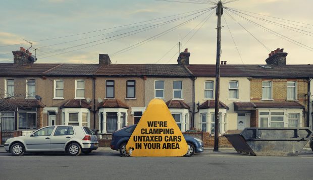 Giant clamp with 'We're clamping untaxed cars in your area' on it on a car in front of row of terraced houses