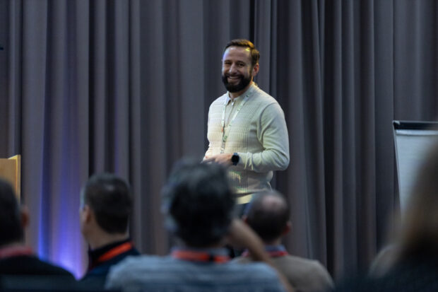 Simon stands in front of a grey curtain giving a presentation, with several audience members seated and facing toward him.