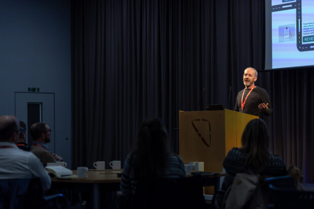 Craig stands behind a podium in a darkened conference room, speaking to an audience at seated tables.