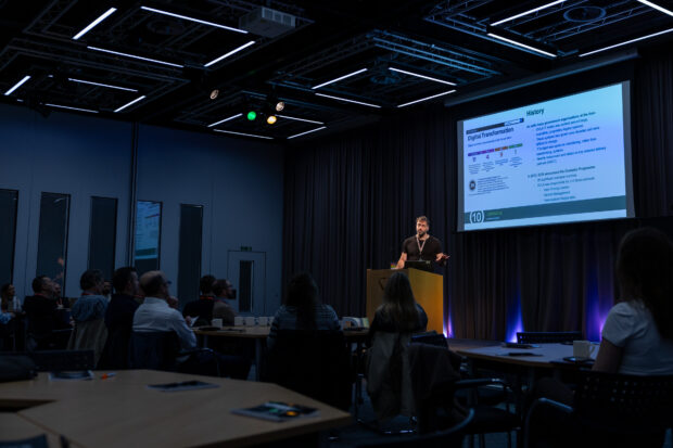 Tom stands at a podium in a dimly lit conference room, presenting slides to an audience. The projected slide displays a timeline.