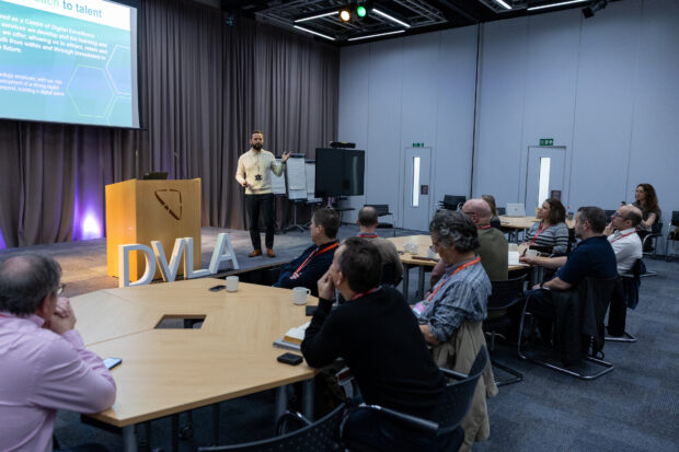 Simon stood at the front of a conference room, presenting to an audience sat at round tables. Large 'DVLA' letters are near the stage, and a presentation slide is projected on the wall behind Simon.